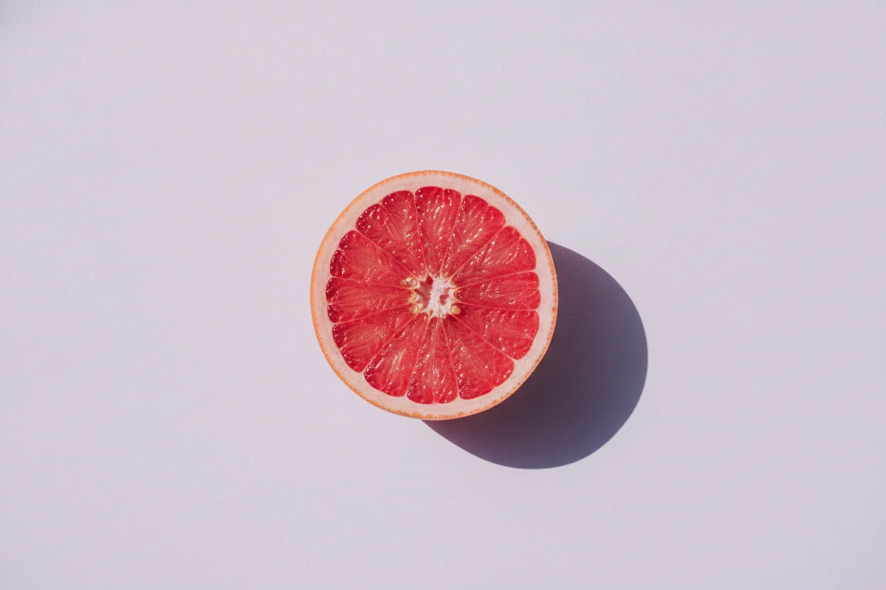A halved pink grapefruit with its cut face upward on a smooth lavender background, lit by harsh flash with a sharp shadow