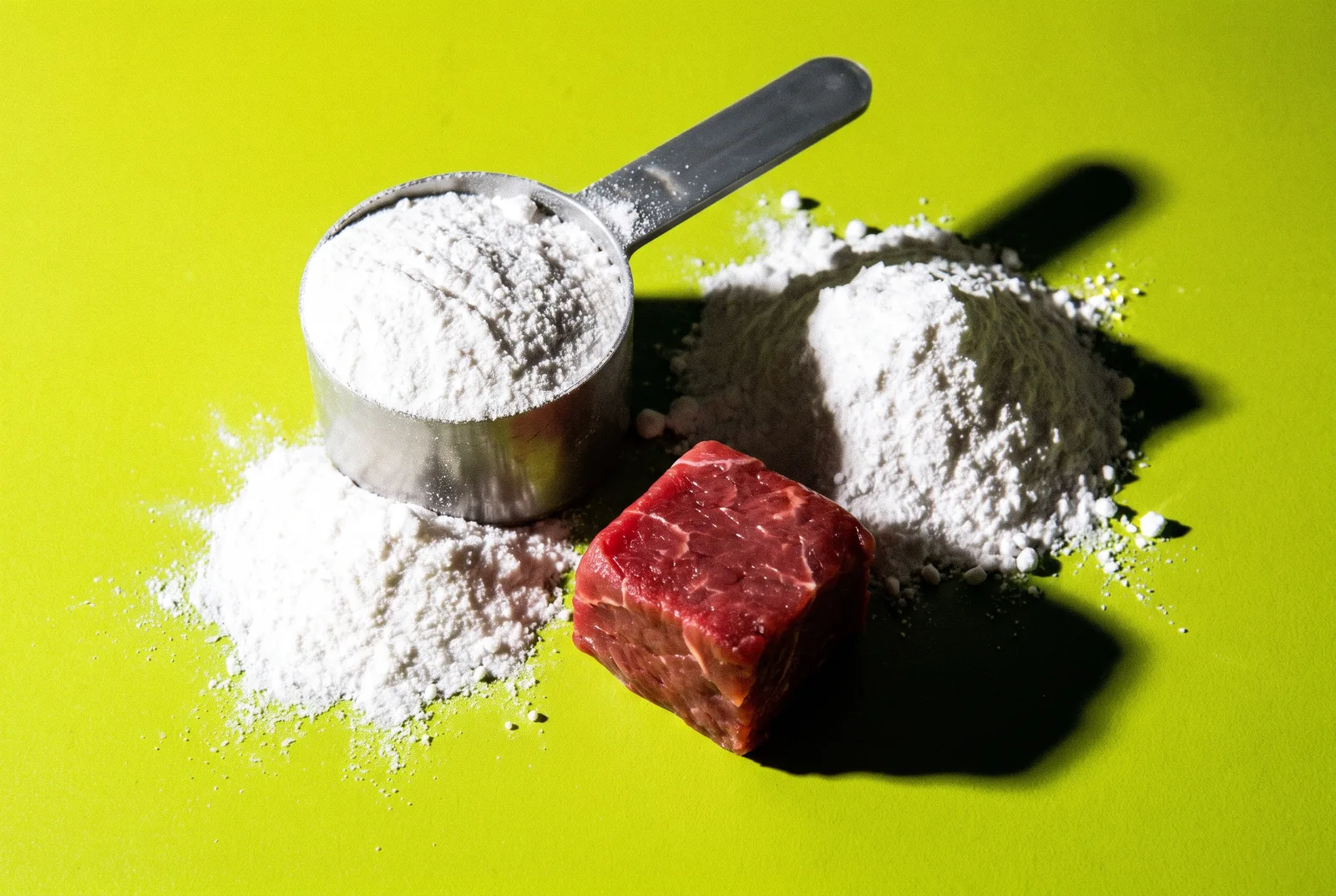 Metal scoop of white creatine powder beside a small spilled mound and a cube of raw red beef on bright lime green