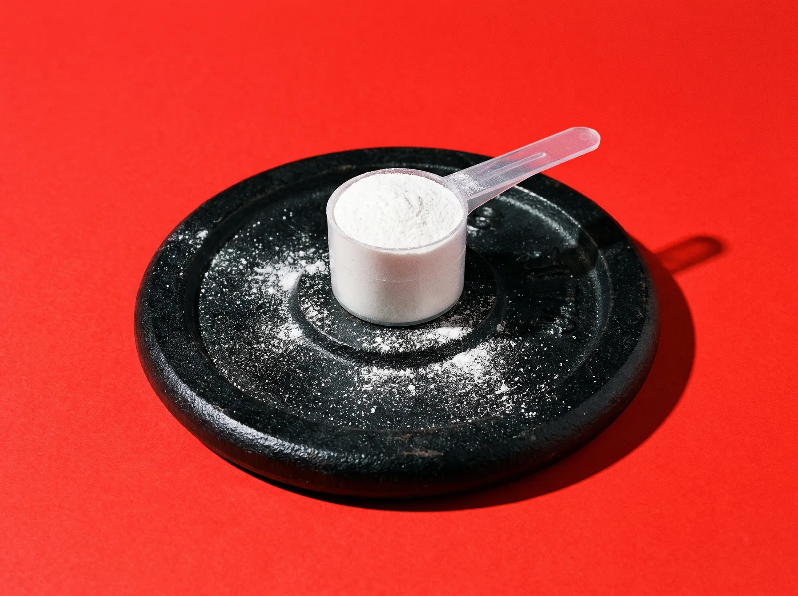 A scoop of white creatine powder on a black weight plate against a red background