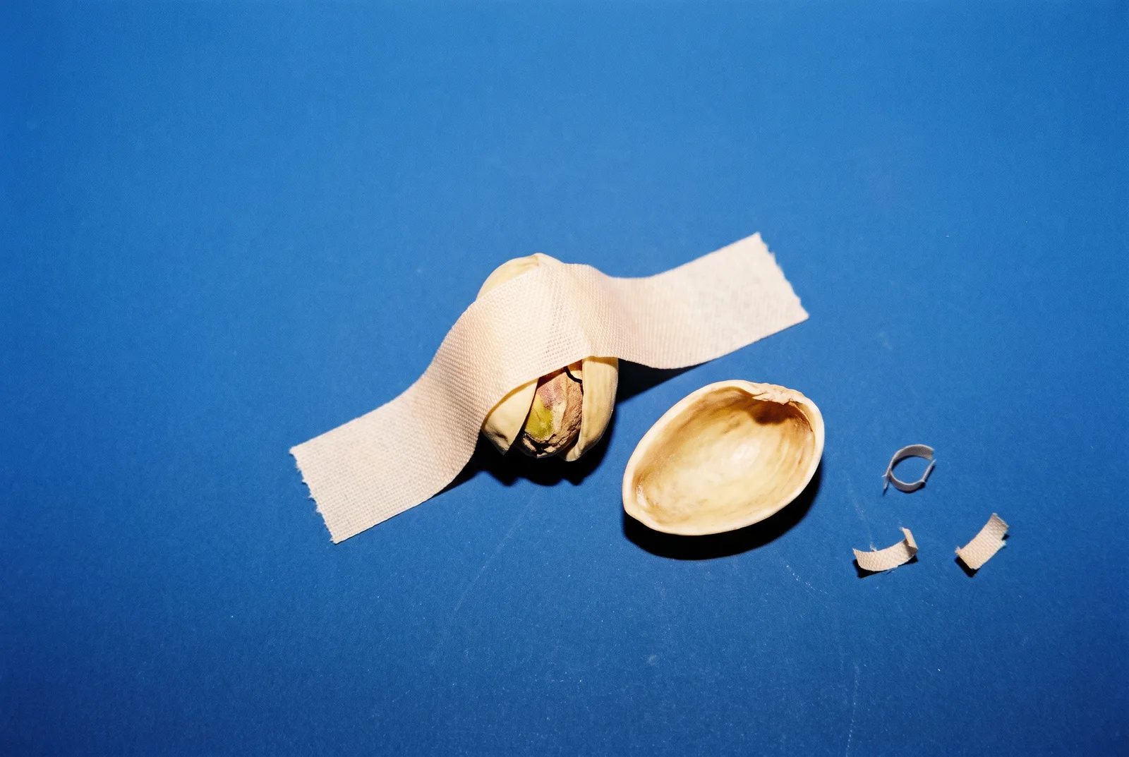 A beige tape strip sealing a pistachio shell shut beside an empty open shell on a bright cobalt blue backdrop