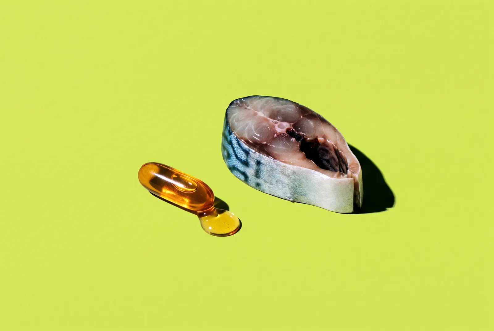 A punctured amber fish oil softgel with a dripping golden droplet beside a raw mackerel steak on a bright lime green background