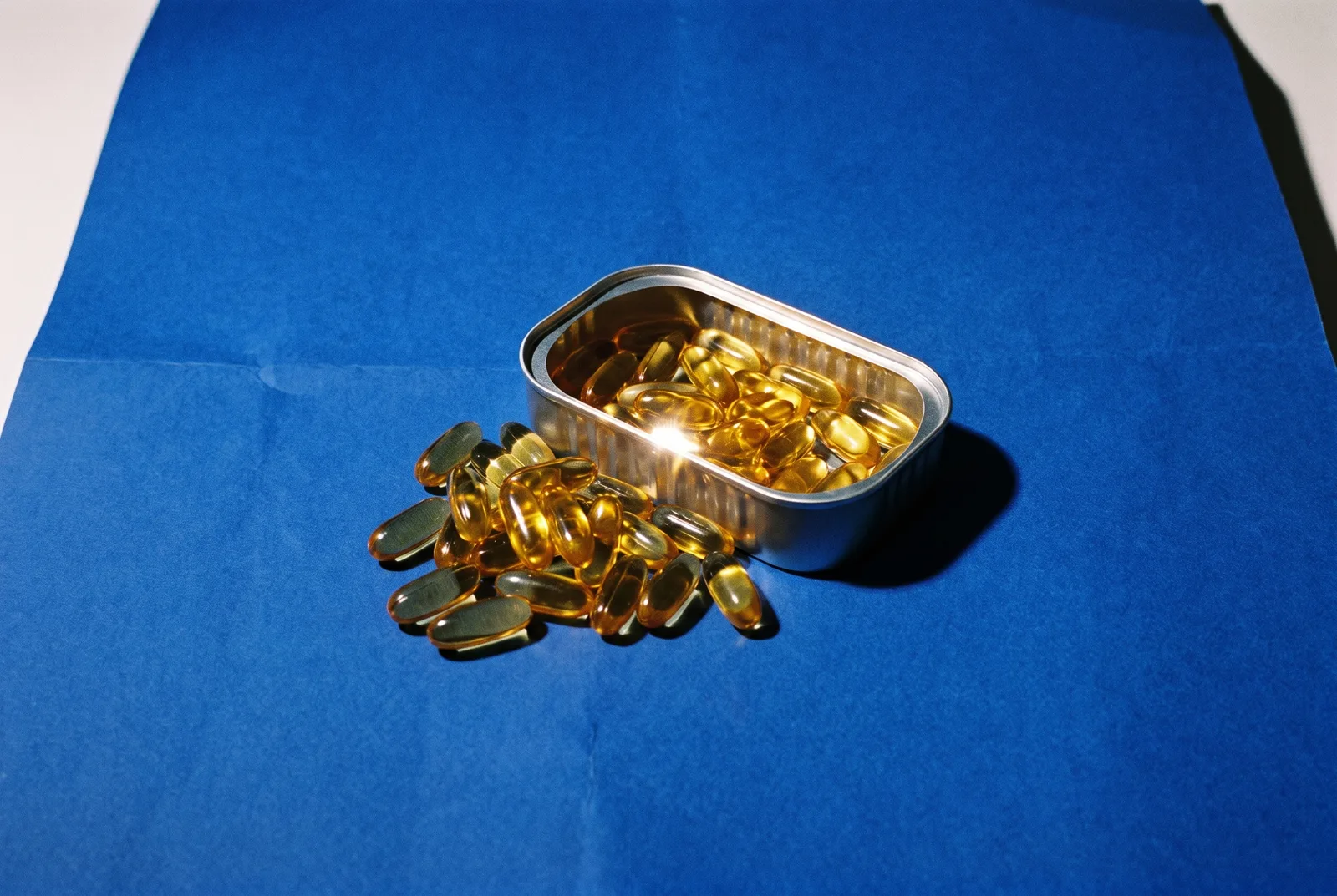 Amber fish-oil softgels spilling from an open sardine tin on a blue background