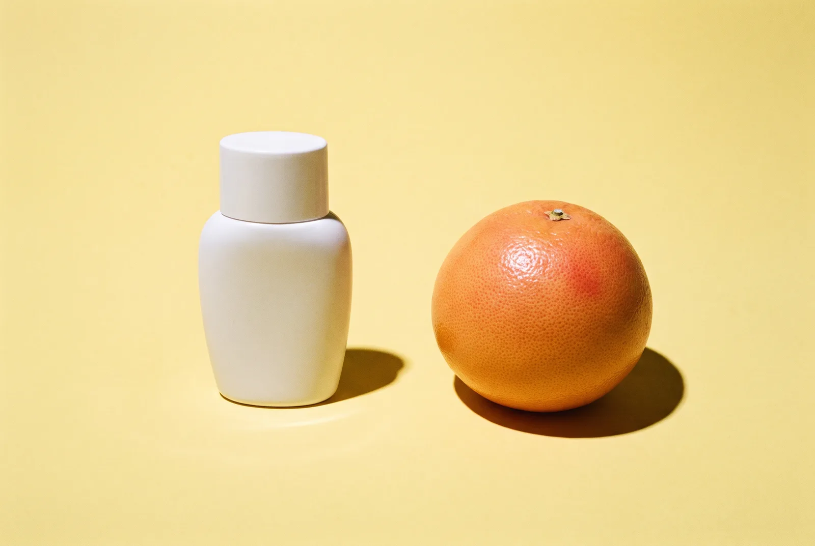 A white sunscreen bottle and a whole pink grapefruit stand apart on a butter-yellow surface under harsh flash with sharp shadows.