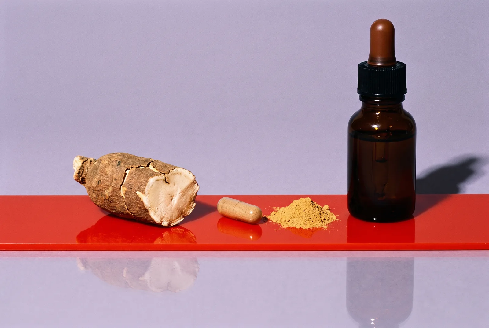 Ashwagandha root, a capsule, a mound of ochre powder and a dark tincture bottle arranged on a glossy red strip against lilac-gray paper