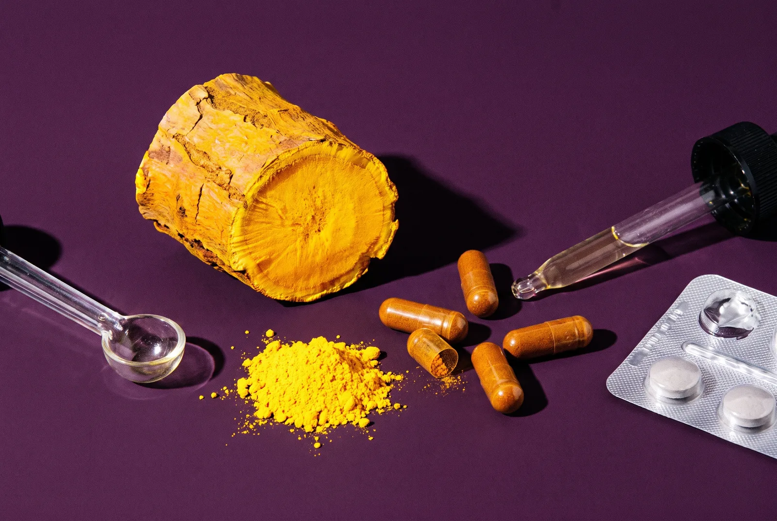 Split yellow berberine root bark, a mound of yellow powder, capsules and a partly empty blister strip on deep plum paper under hard flash