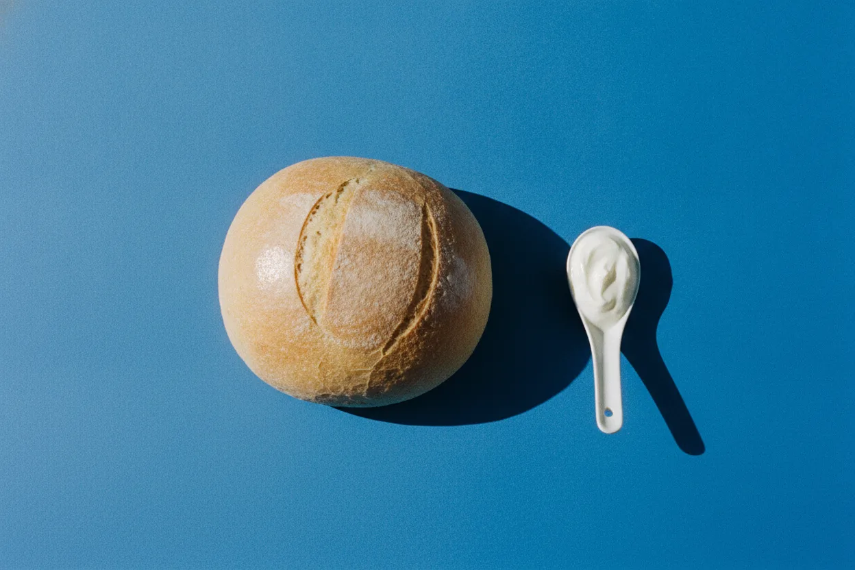 A round loaf of bread and a small spoon of white yogurt on a bright cobalt-blue background under harsh flash