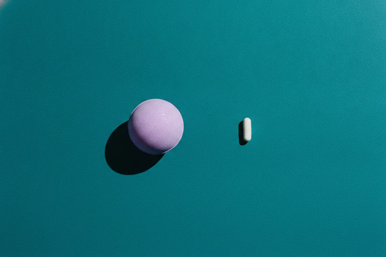 A lavender-purple bath bomb sphere and a small white capsule sit apart on a deep teal paper background under hard flash.