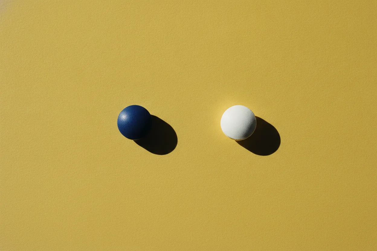 A navy blue round pill and a white round pill lie separately on a bright mustard yellow surface with sharp shadows