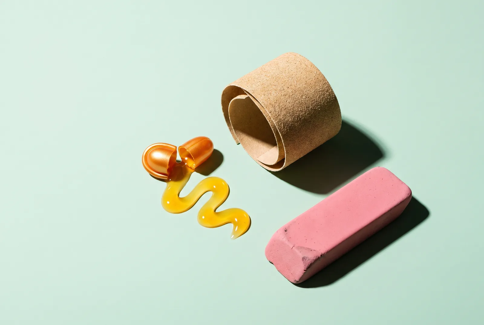 An opened orange retinol capsule with a spiral of glossy serum beside a sandpaper strip and a worn pink eraser on pale mint